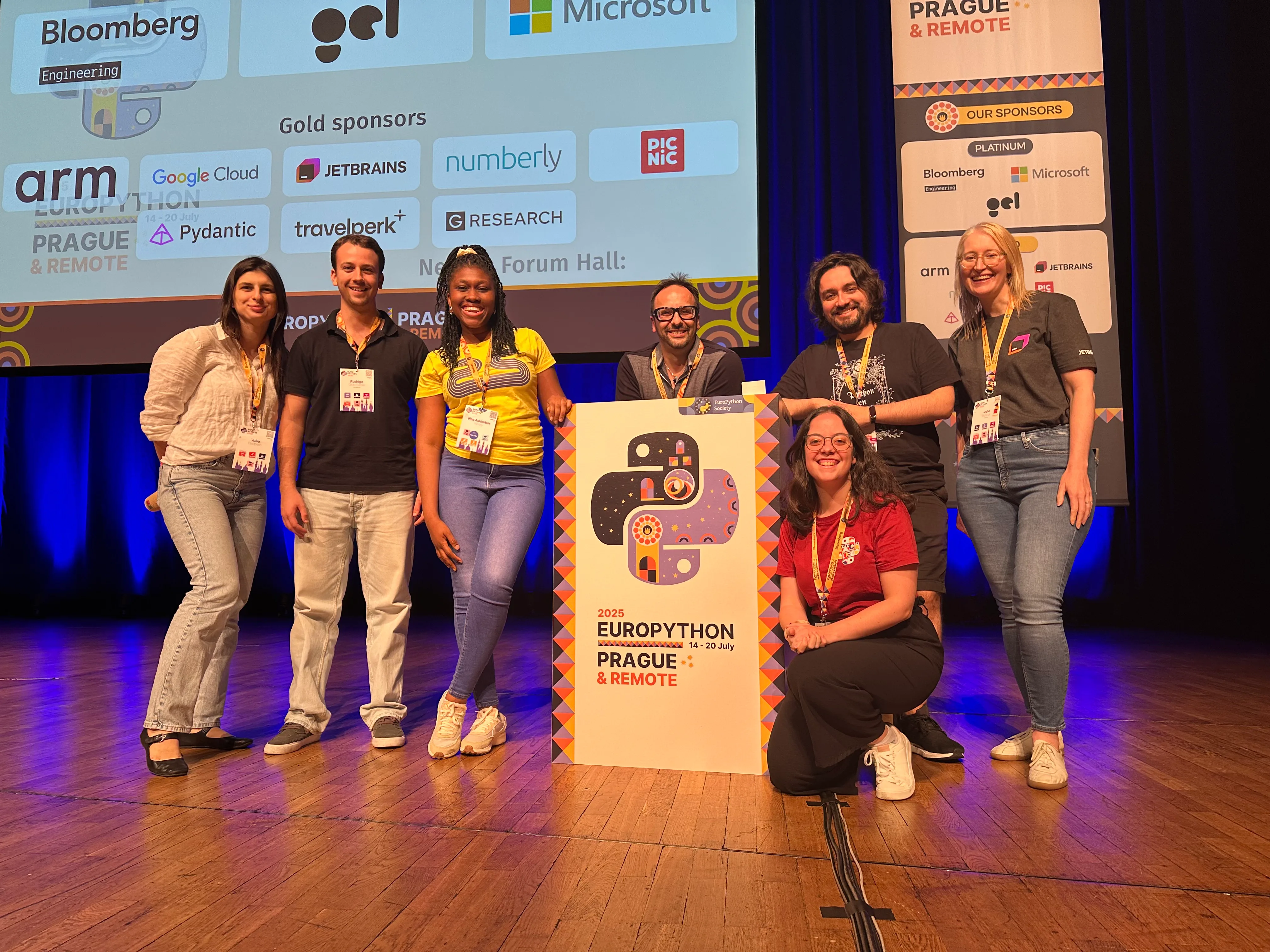 A picture of the EuroPython 2025 programme team in the main stage surrounding the speaker podium. From left to right, Yuliia, Rodrigo, and Naa, are to the left of the podium; Diego is behind it; Cristián and Jodie are to the right of it; and Marina is kneeling next to the podium in front of Cristián. Everyone is smiling.