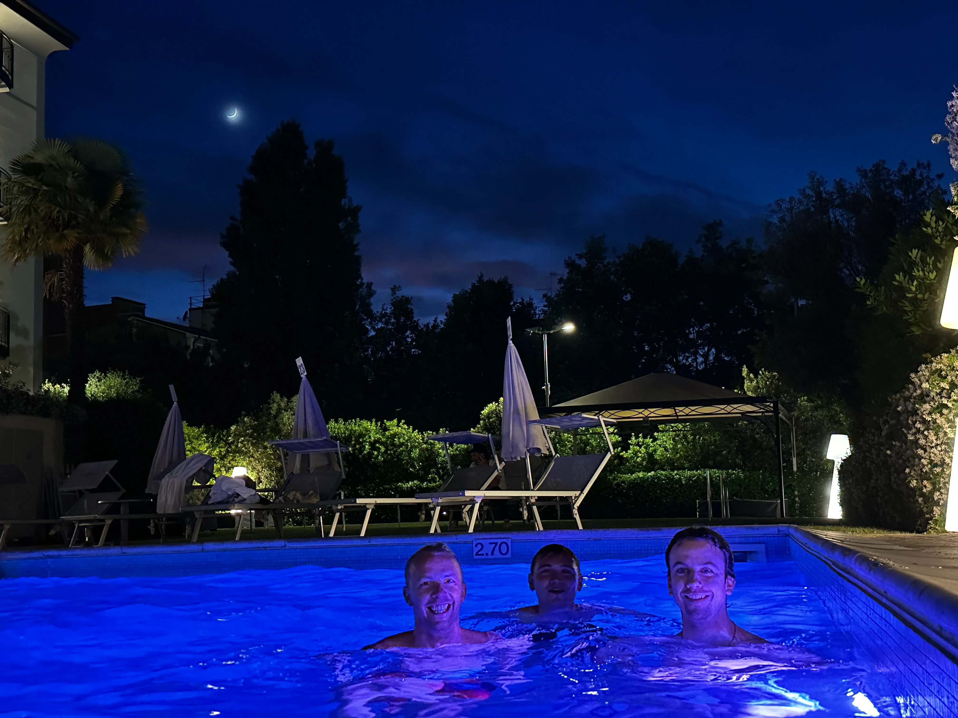 A dark picture of a pool by night with three people in it more or less smiling at the camera.