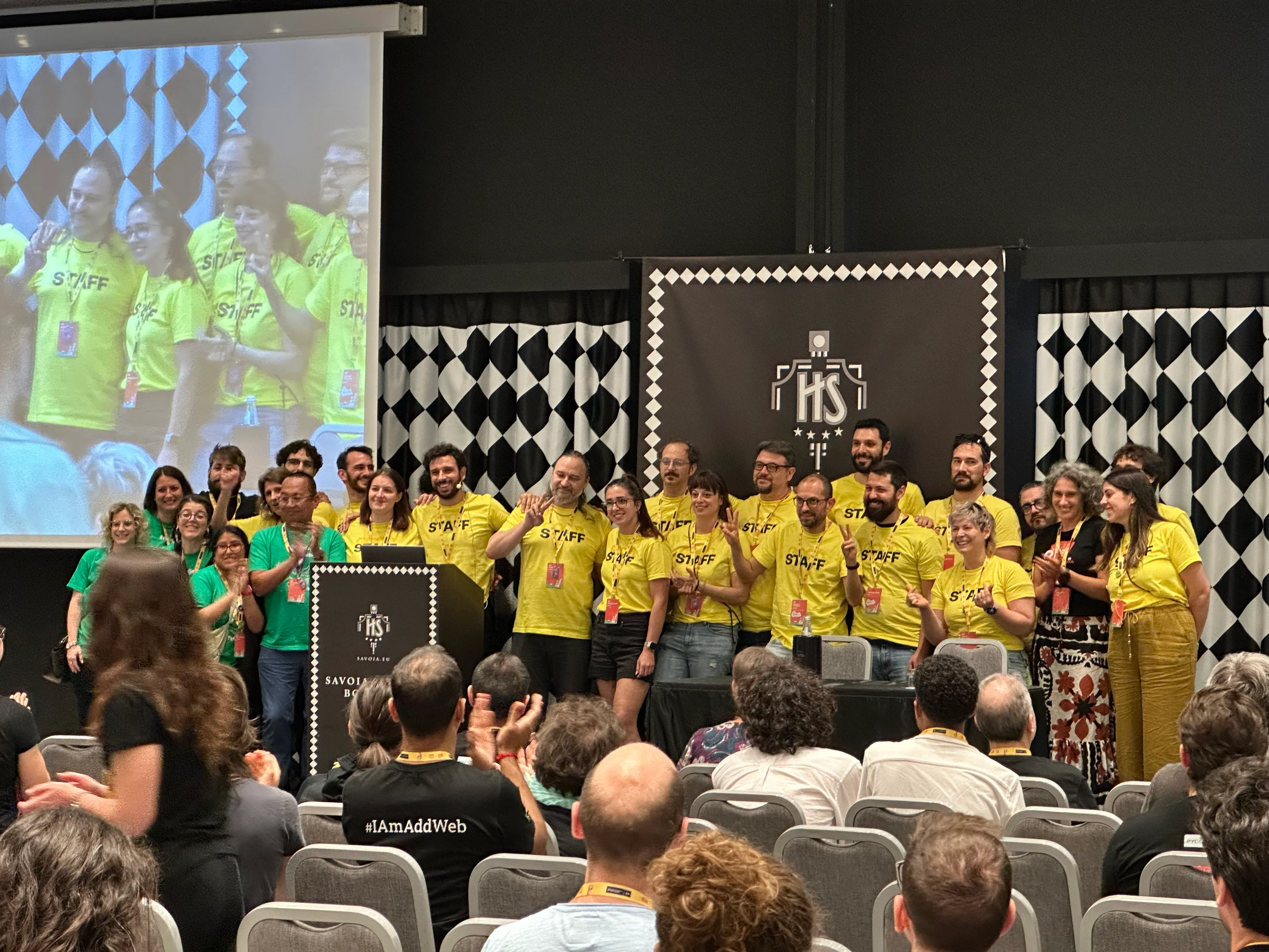 A photo of dozens of PyCon Italy volunteers on stage being applauded for their phenomenal work. Most volunteers are wearing the same yellow t-shirt but a small group on the left is wearing left t-shirts.