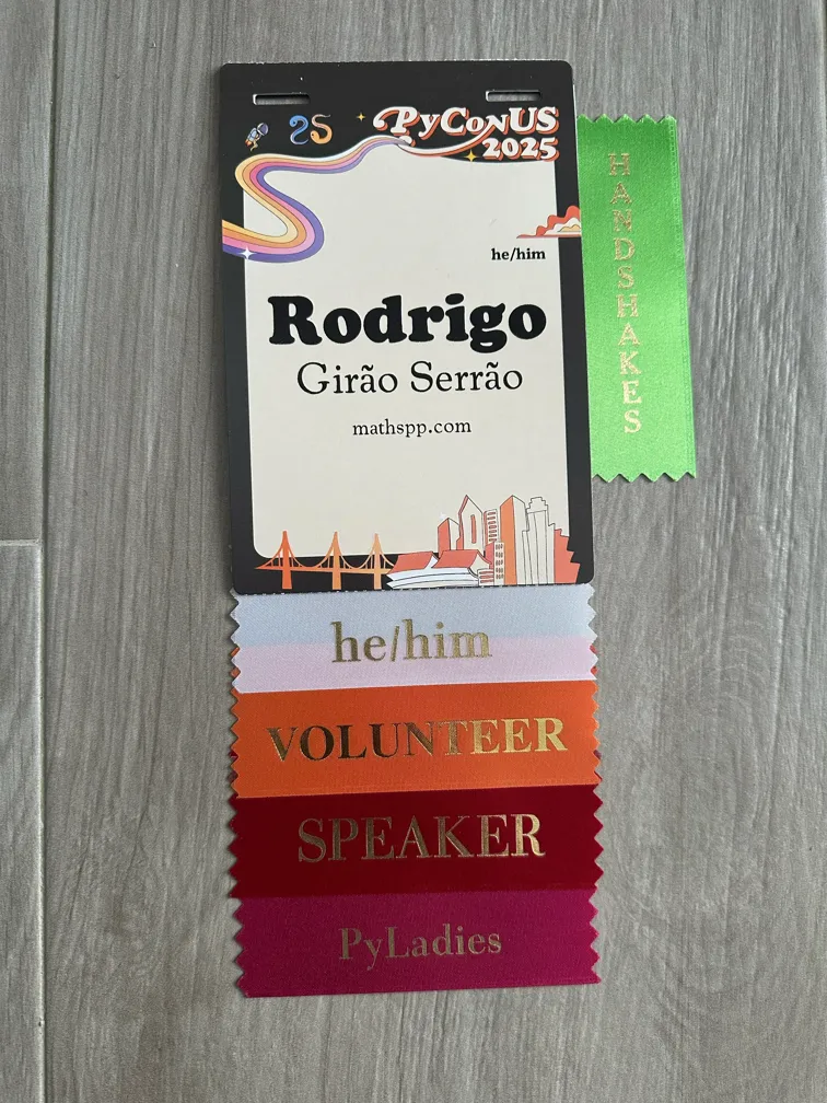 My badge for PyCon US, showing my name and some ribbons I attached: he/him, pyladies, speaker, volunteer, handshakes