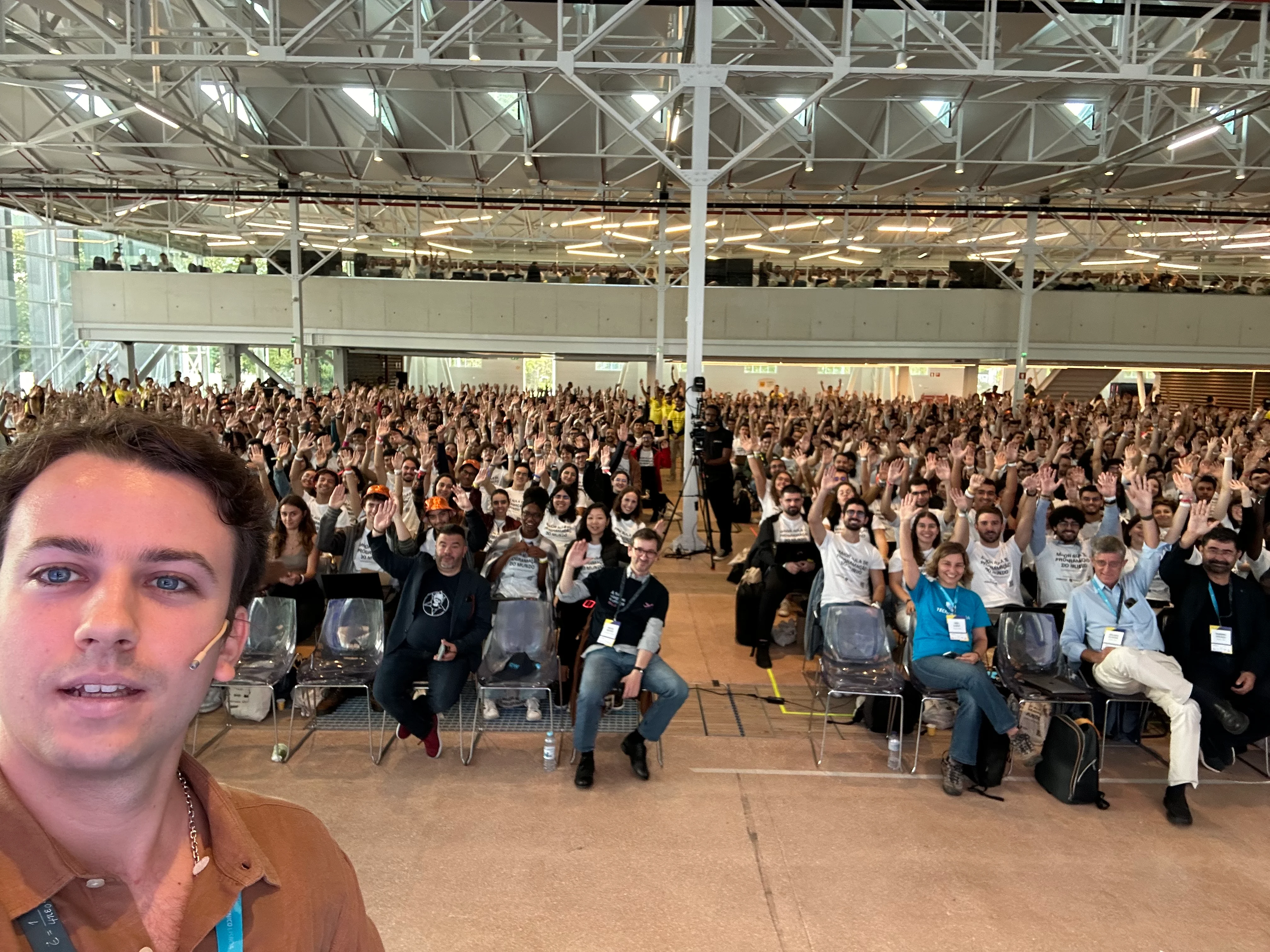 Selfie showing a large room with a sea of people that goes so far back that you cannot really count how many rows of chairs there are. You cannot see all of the students who were trying to break the Guinness World Record for the largest programming lesson. Everyone has their arms up and part of my face is visible on the left.